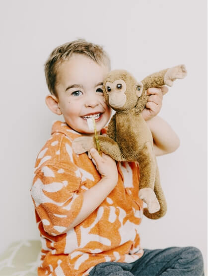 EIn kleiner Junge hält fröhlich lachend seinen Kuscheltier-Affen und eine Zahnbürste hoch